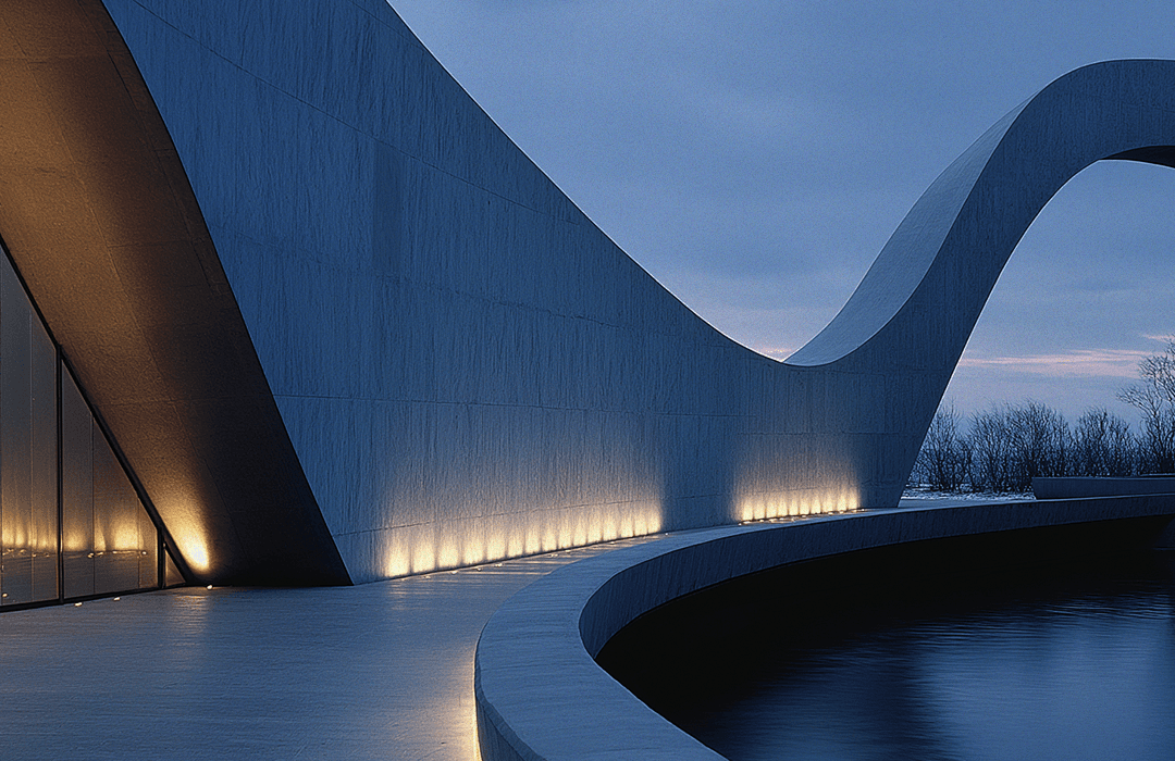 Vista de perto e externa de uma arquitetura moderna e orgânica. A estrutura é uma grande parede de concreto cinza ou azul-escuro com uma forma curvilínea, suave e ondulada, que se eleva e se curva em direção ao céu, lembrando uma onda ou uma letra 'S' esticada. Na base da parede, há uma linha de luzes quentes (uplights) iluminando a superfície e criando um contraste dramático com o céu crepuscular azul-acinzentado. Em primeiro plano, há um caminho curvo sobre uma superfície plana clara e, à direita, um espelho d'água escuro, que acompanha a curvatura da parede. Ao fundo, árvores sem folhas são visíveis sob a luz fraca. A imagem evoca uma sensação de monumentalidade e calma no cair da noite.