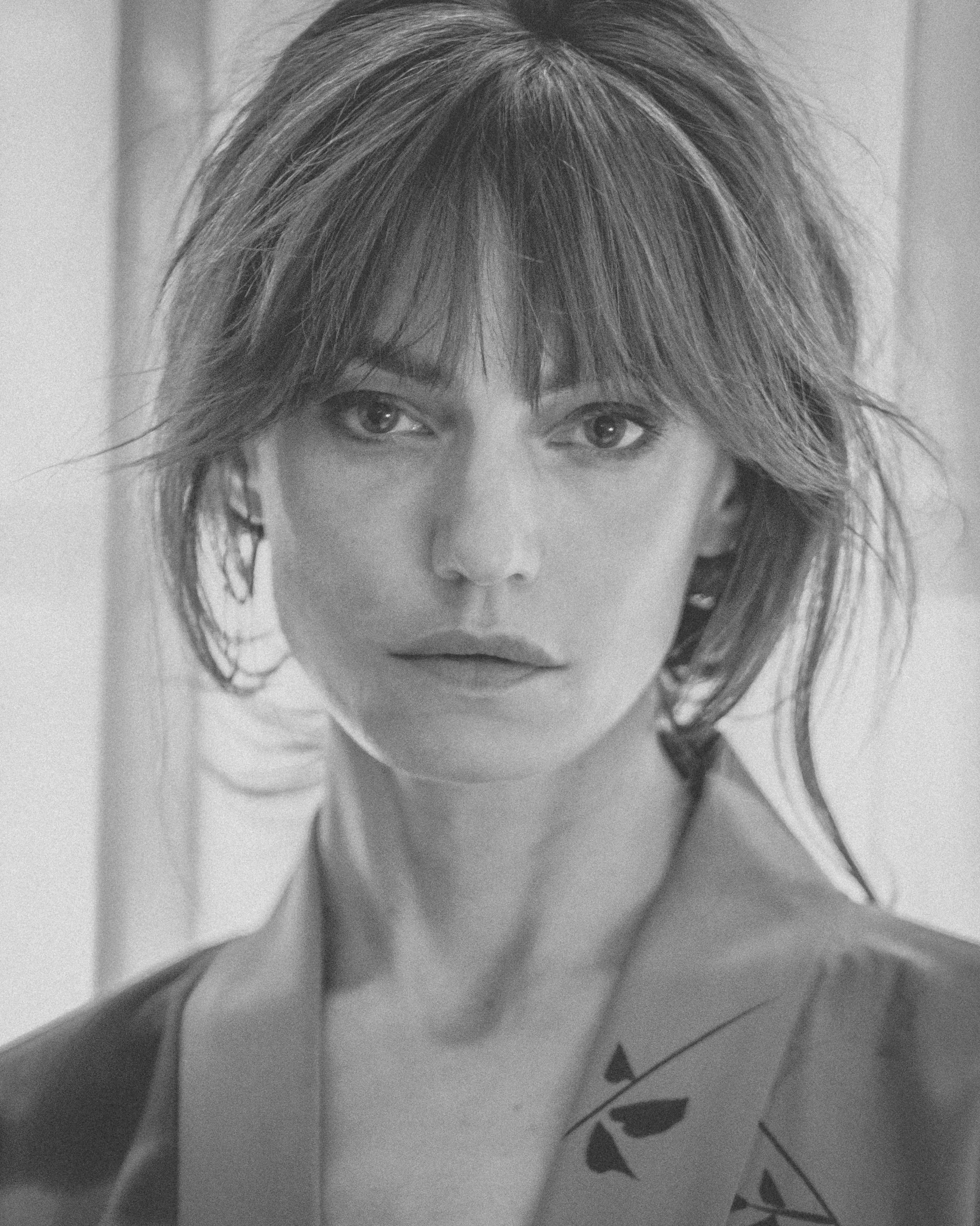 Black-and-white close-up of a woman with wispy bangs and an open blazer, soft backlight through curtains.