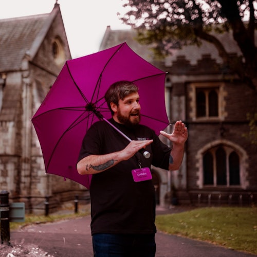 一位留着胡子的男子手持粉色雨伞，在历史建筑和树木附近的户外比划着并交谈。