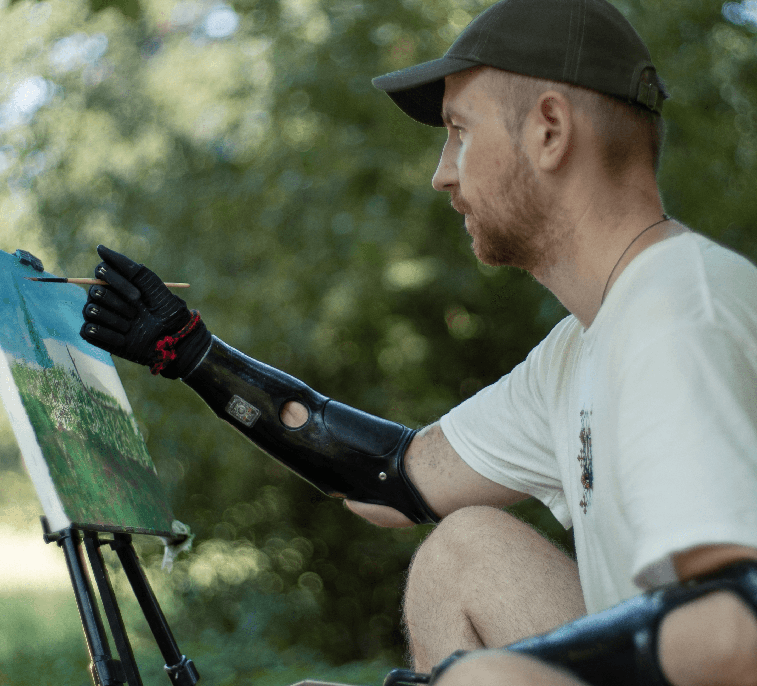 A man with dual black prosthetic arms Esper Hand sits on the grass painting a canvas on an easel in a green park, capturing a serene landscape.