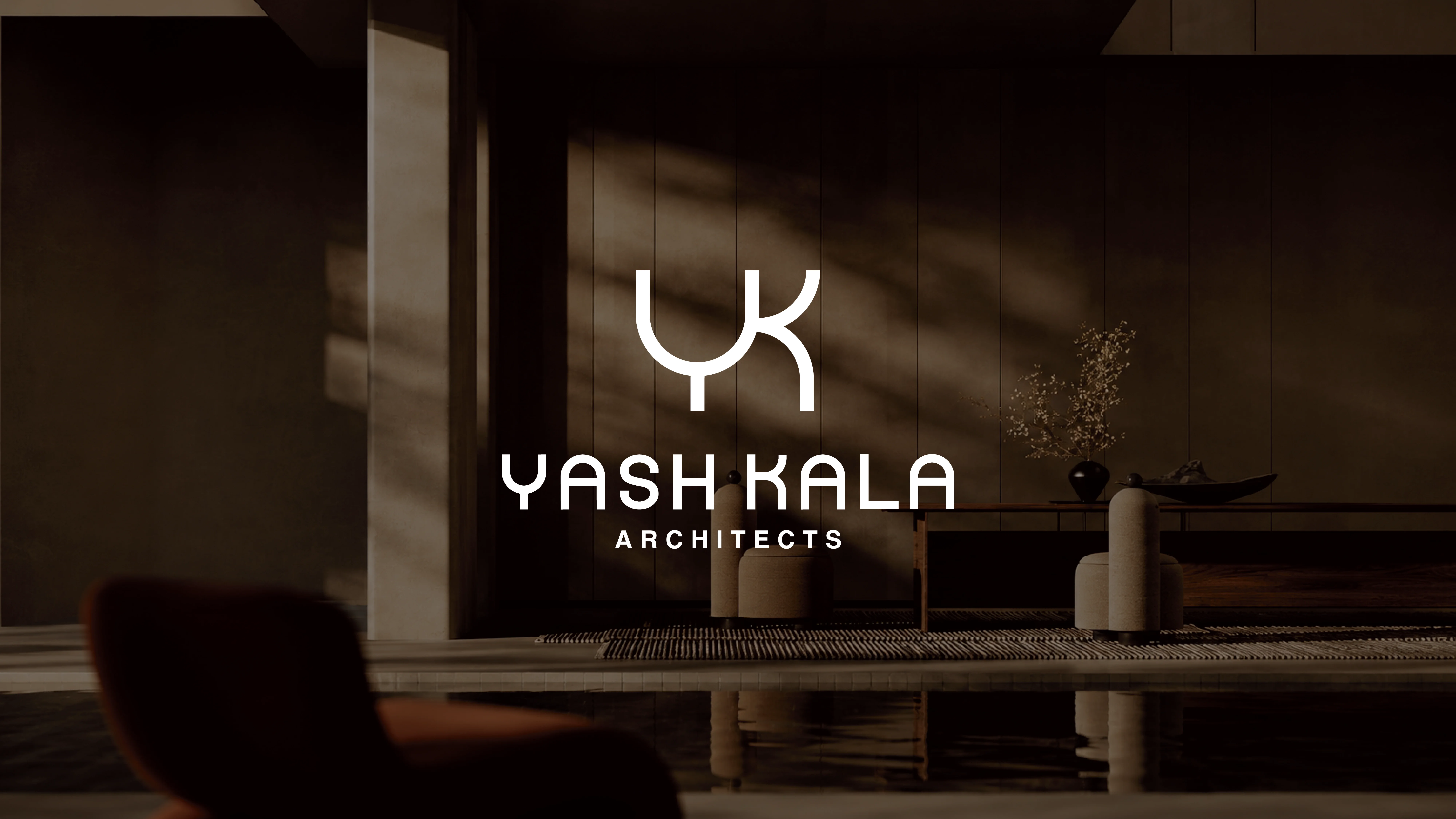 Modern architectural interior of a dimly lit room with minimalist furniture, featuring a reflective floor, subtle shadows, and the logo "Yash Kala Architects" prominently displayed on a wall.