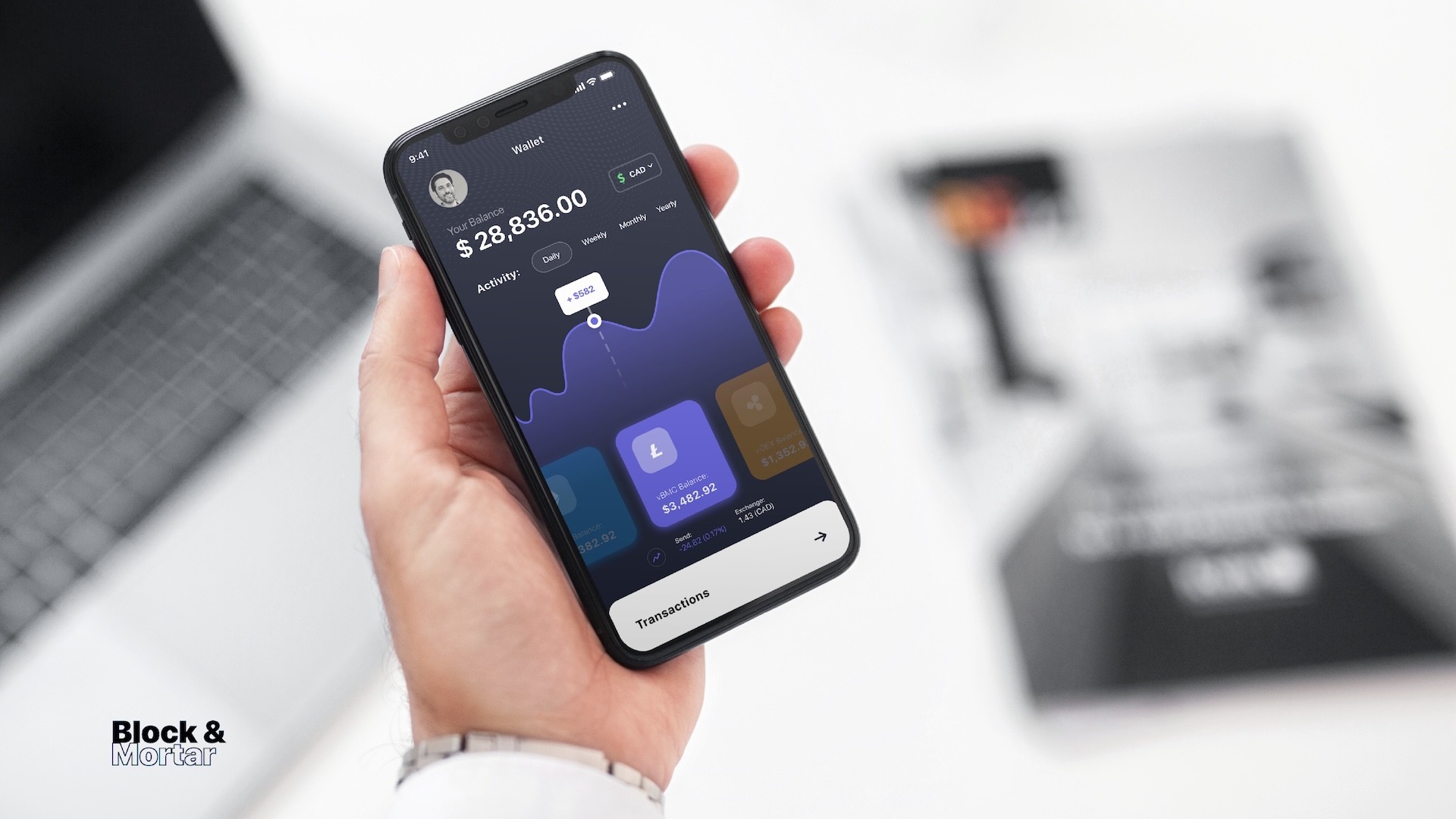 Lifestyle mockup of Block & Mortar wallet screen on iPhone held in hand at desk, displaying $28,836 balance with purple activity graph, Litecoin currency card selected showing $3,482.92, demonstrating multi-currency blockchain wallet UX design