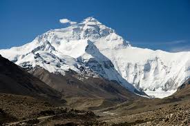 A wide, majestic view of a massive, snow-capped mountain, likely Mount Everest, under a bright blue sky. The lower slopes are rocky and brown, leading up to the towering white peak and dramatic ridgelines covered in glaciers and deep snow.