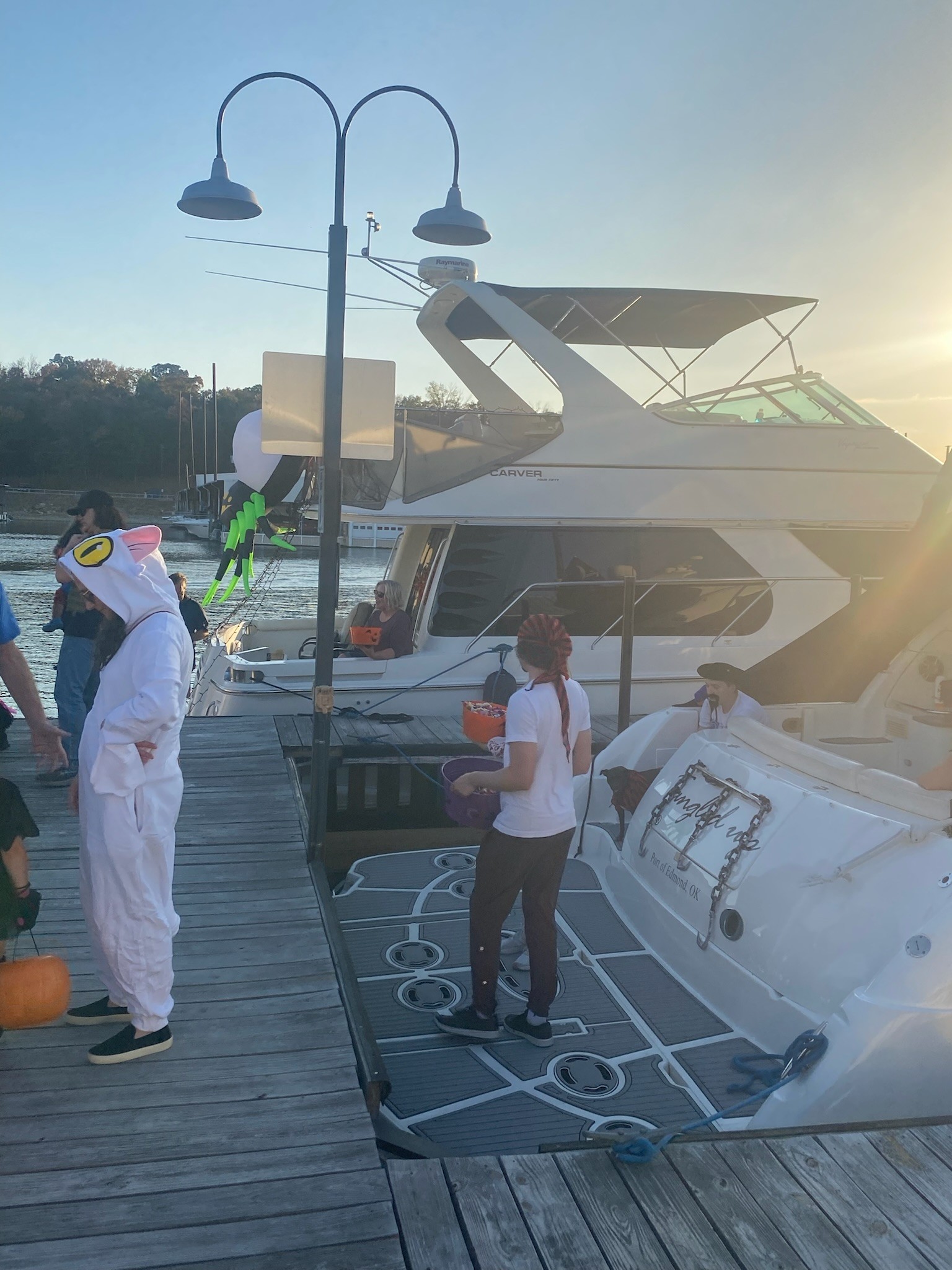 A group of people dressed in costumes, including a penguin and a pirate, stand on a dock beside a yacht with the sun setting in the background.