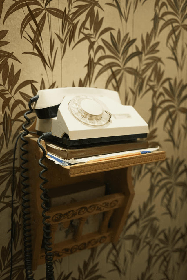 Vintage rotary telephone placed on a small wooden shelf, with papers stacked underneath and patterned wallpaper in the background.