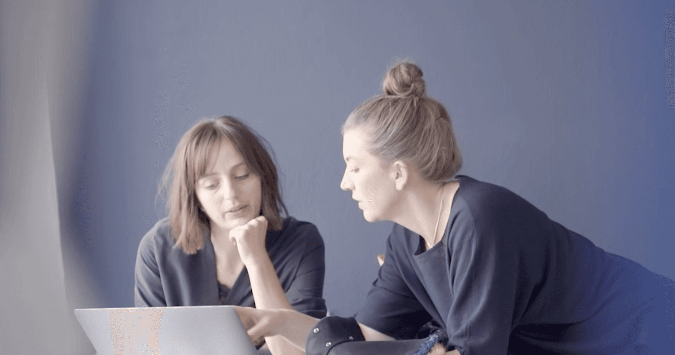Two women are sitting and discussing something on a laptop screen against a neutral gray background, conveying a sense of collaboration and teamwork.