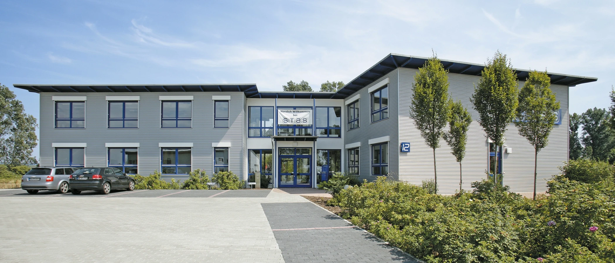 Modern office building with corrugated metal siding, blue-framed windows, a glass entrance, and surrounding greenery.