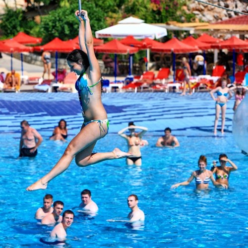 A woman in a bikini is zip-lining over a crowded pool, with people swimming and sunbathing under red umbrellas in the background.