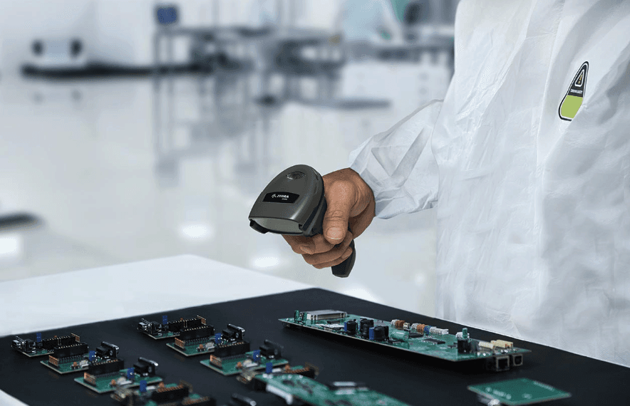 A person in a white lab coat holds a handheld barcode scanner over a workbench with multiple electronic circuit boards, set in a clean, softly lit lab environment.