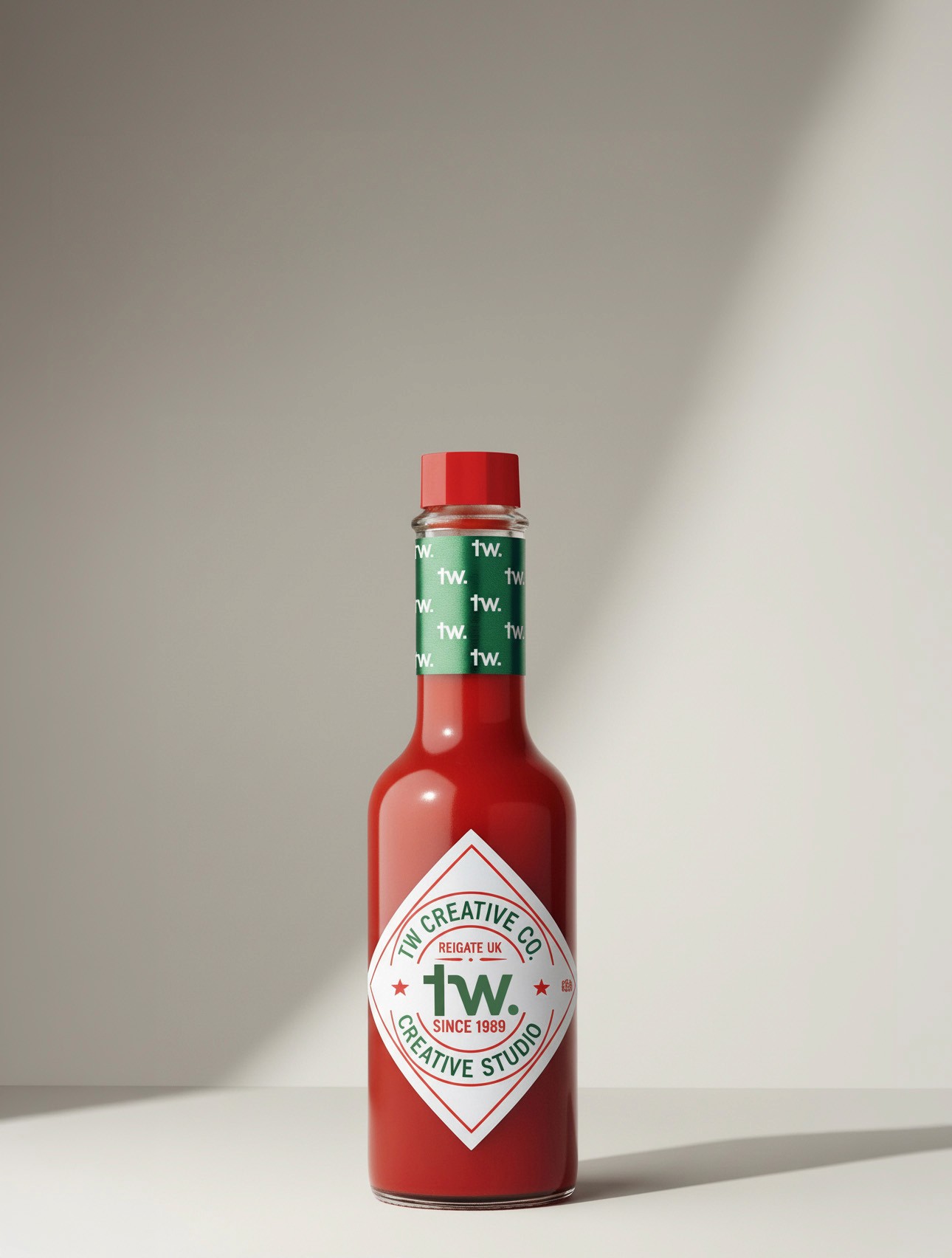 A red hot sauce bottle on a clean neutral background, featuring a custom TW Creative label and a green neck band patterned with the “tw.” mark.