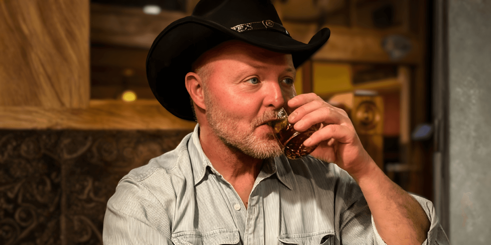 A man wearing a cowboy hat enjoys a drink, capturing a moment of calm and reflection in a rustic setting.