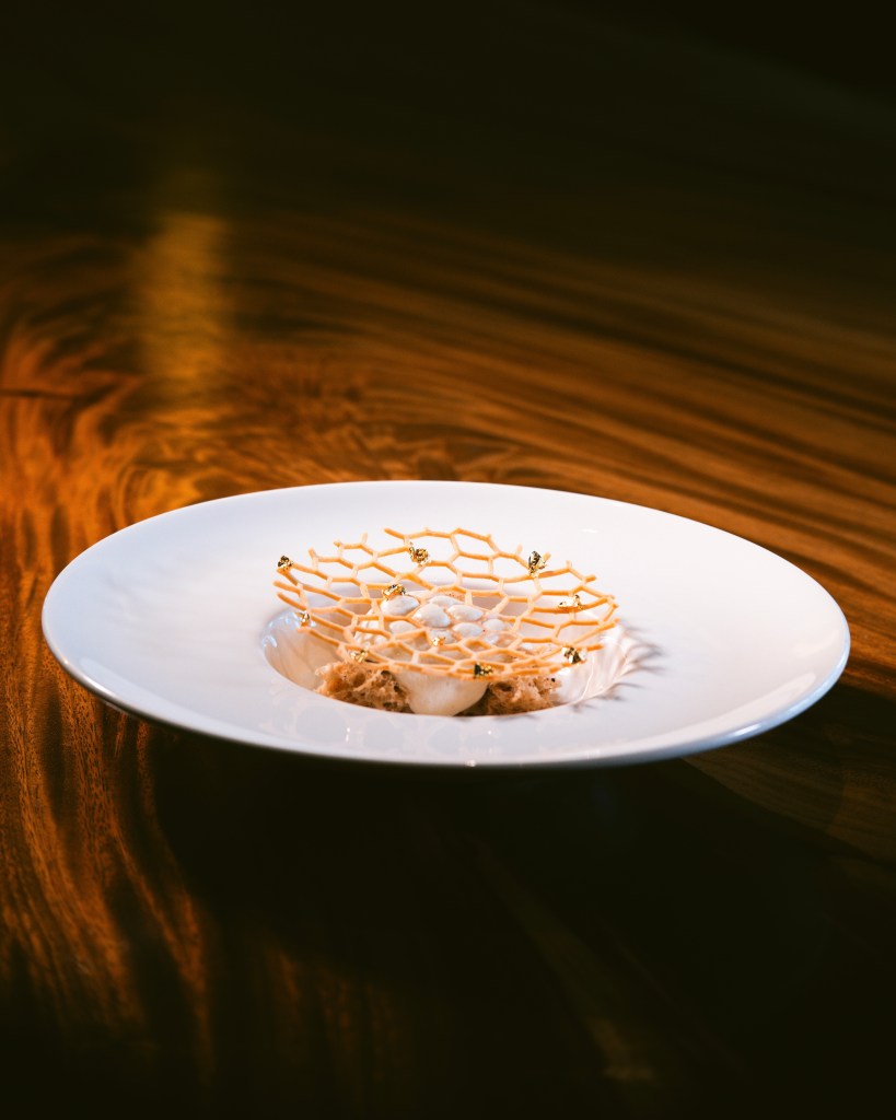 A beautifully presented dessert featuring a honeycomb-like garnish atop a creamy base, adorned with edible gold flakes, set against a warm wooden table.