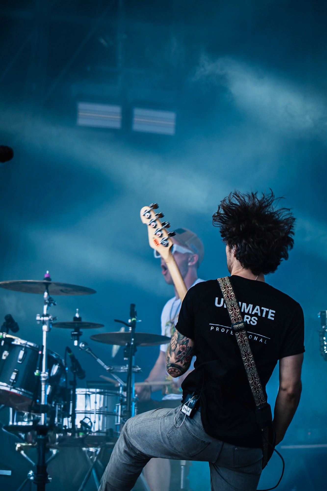 Guitariste de dos, cheveux en mouvement, batteur flou en arrière-plan, lumière bleue