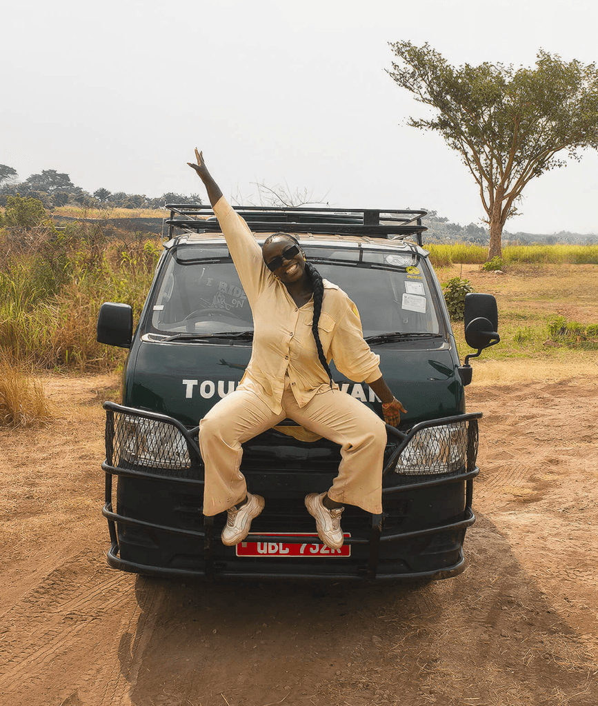 Mary sitting on the bonnet of a car, posing confidently, with an Uganda's landscape in the background.