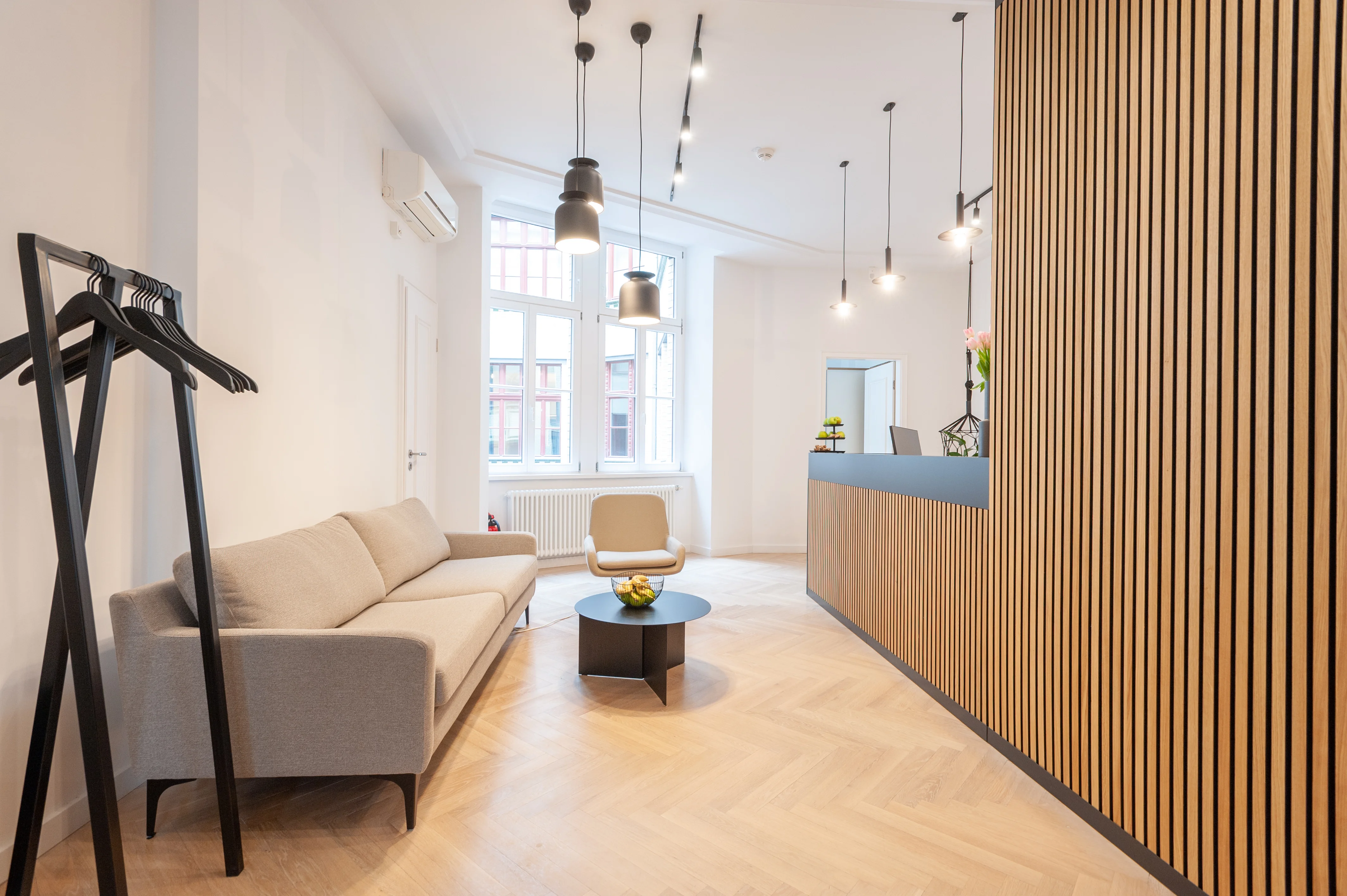 Waiting lounge at AI START-UP BERLIN with a beige sofa, a black round coffee table with fruit bowl, a beige armchair, black coat stand and a reception counter behind a wooden slat wall with petrol-blue top.