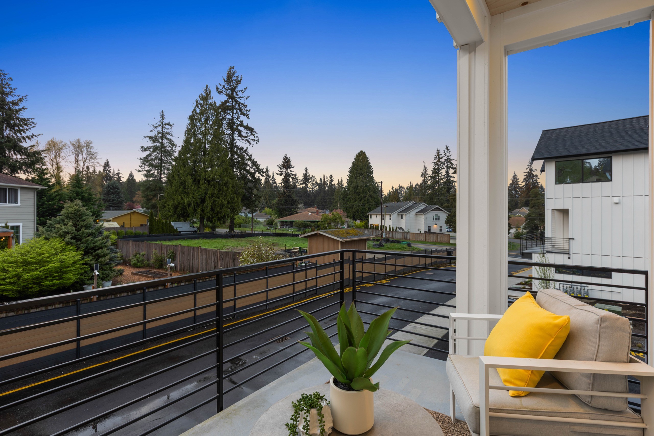 Expansive private balcony of a luxury townhome at dusk with black metal railing, yellow accent cushions, potted plants, and panoramic views of green trees and open sky