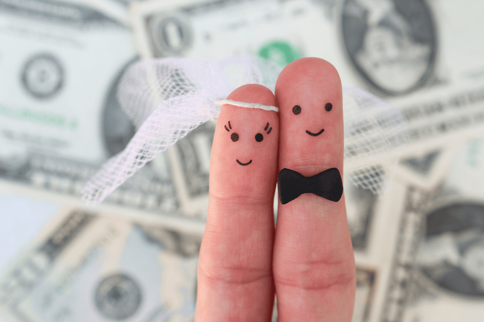 Two fingers dressed as a bride and groom with drawn-on faces, posed against a background of scattered dollar bills.