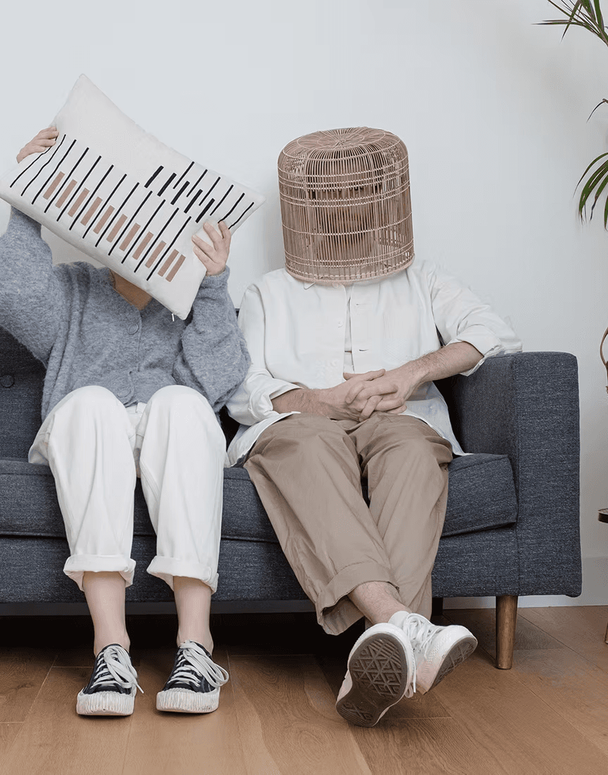 Two people sitting on a blue sofa with one hiding behind a striped pillow and the other with a woven basket on their head, adding a playful and whimsical touch to the living room scene.