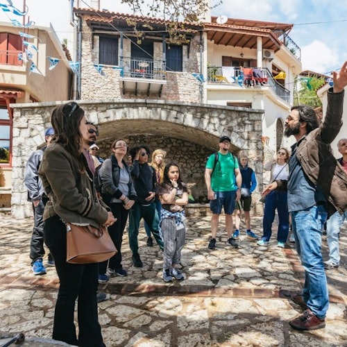 Um guia turístico fala com um grupo de pessoas em um pátio de pedra, com edifícios ao fundo.