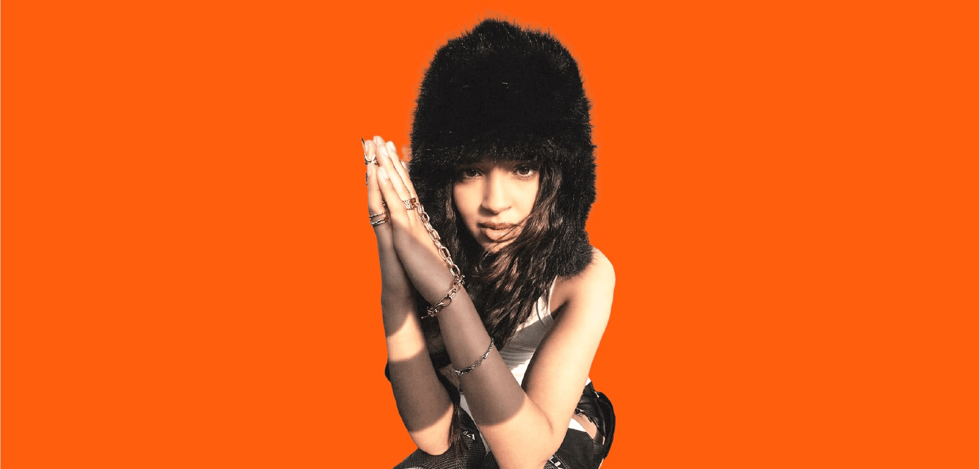 Bannière au format panoramique avec un fond uni orange vif (couleur d'identité de TopLine). À droite, un portrait tronqué et stylisé d'une artiste musicale (rap/pop) ou d'une influenceuse. Elle porte un bonnet en fausse fourrure noire et de multiples bagues et chaînes dorées. Ses mains sont jointes devant son visage, créant une pose accrocheuse. L'espace négatif à gauche est dédié au texte. Ce visuel de direction artistique forte est utilisé pour l'identité visuelle et le branding du média digital musical TopLine, mettant en évidence les services de graphisme et de création de contenu de Nux-Studio.