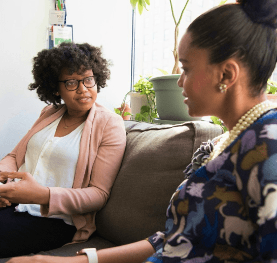 Two women sitting on a couch in a bright room, engaged in conversation.