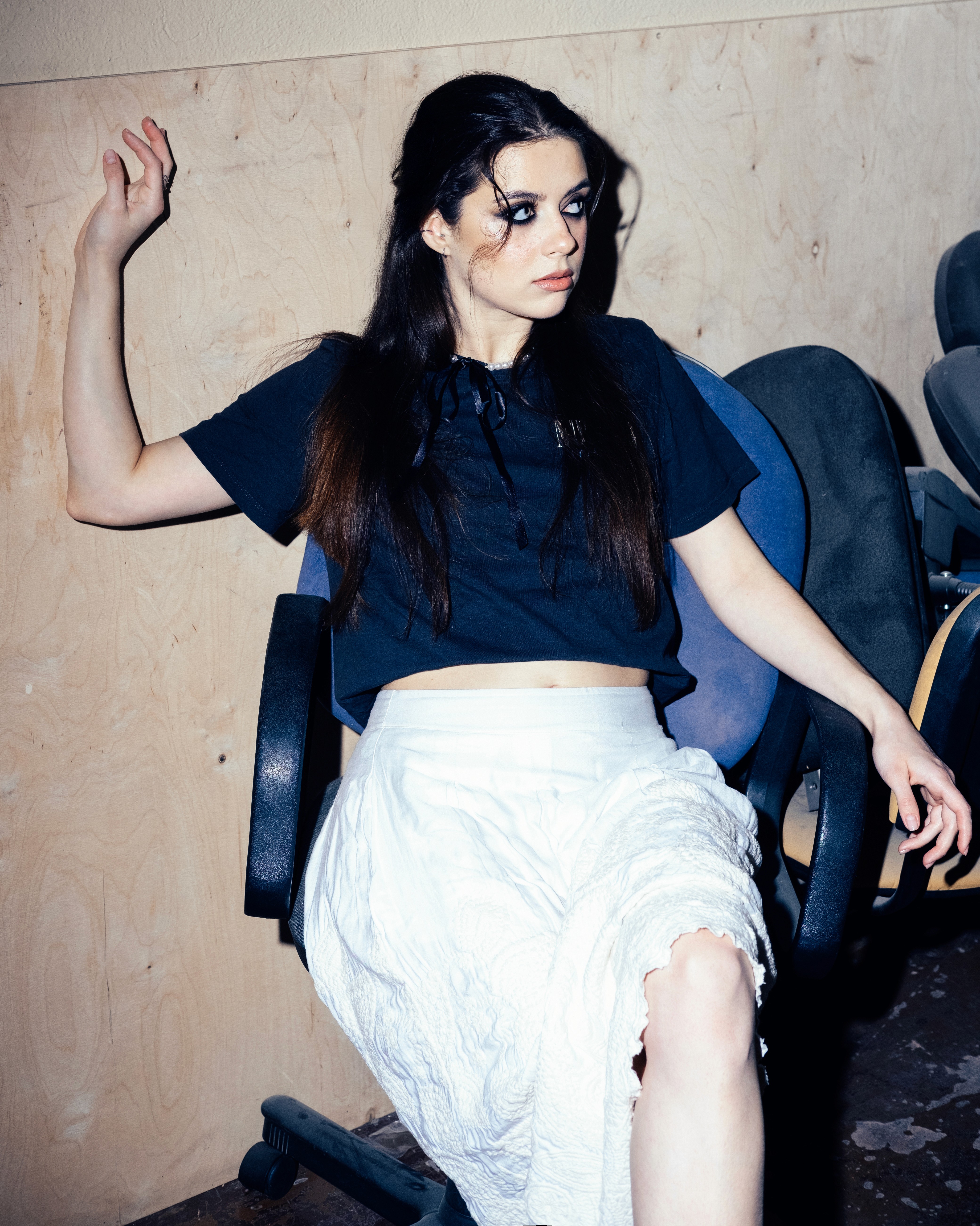 Tanya – Youth Disorder – seated on a chair in a sparse room, arm lifted with a distant expression