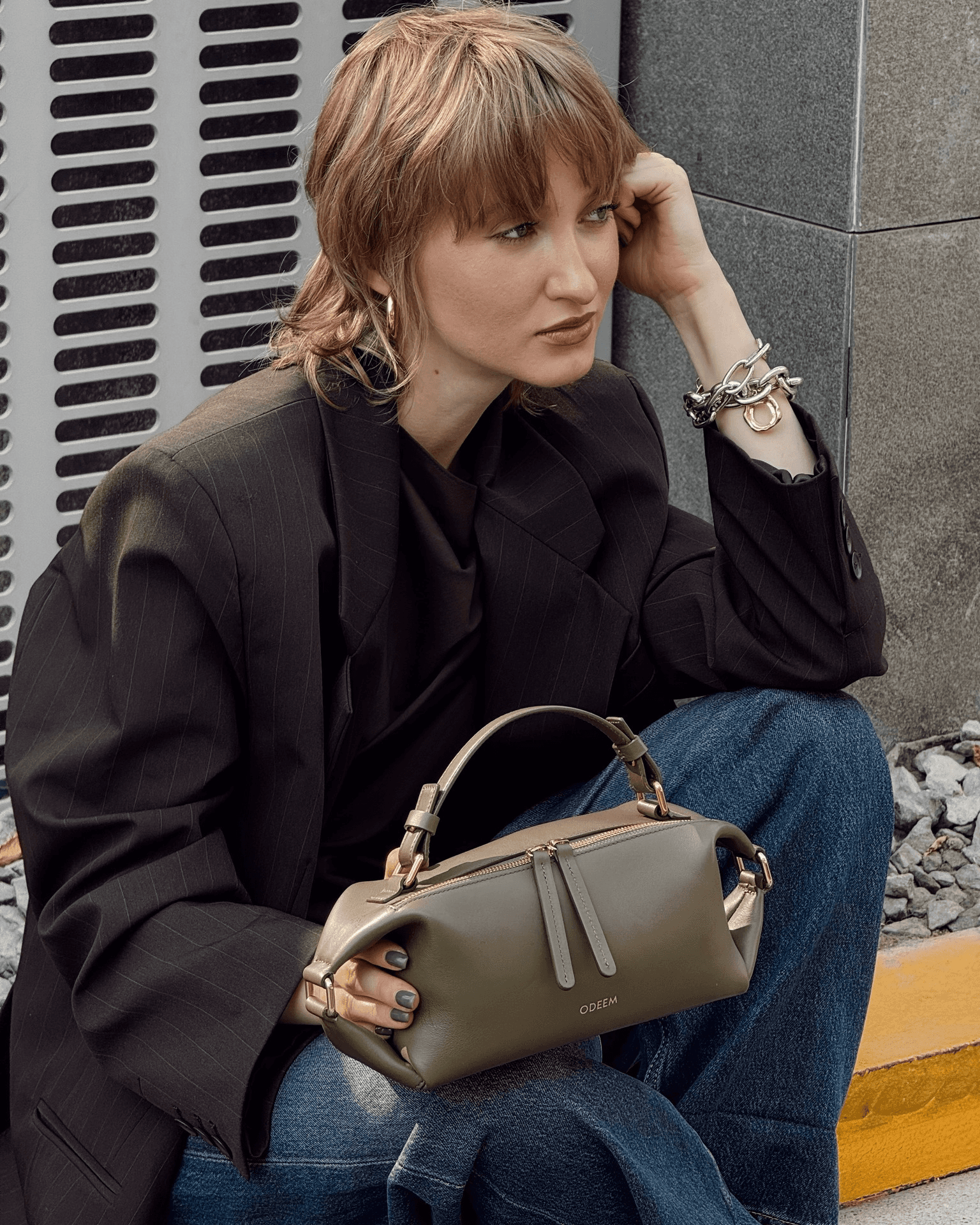 Close-up of a large, structured brown leather tote bag held by a person in a belted khaki trench coat, shown against a textured natural backdrop.