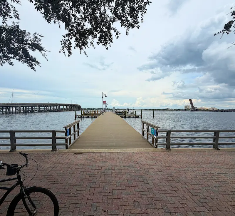 A serene view of the Riverwalk in Bradenton, FL, featuring a long wooden pier extending into calm waters, flanked by railing, with cloudy skies above and a bridge visible in the distance near the Gulf Islands Ferry stop.