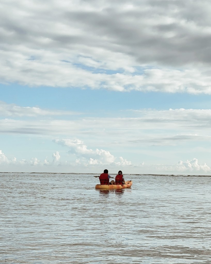 Two people in life jackets kayaking in calm water under a partly cloudy sky.