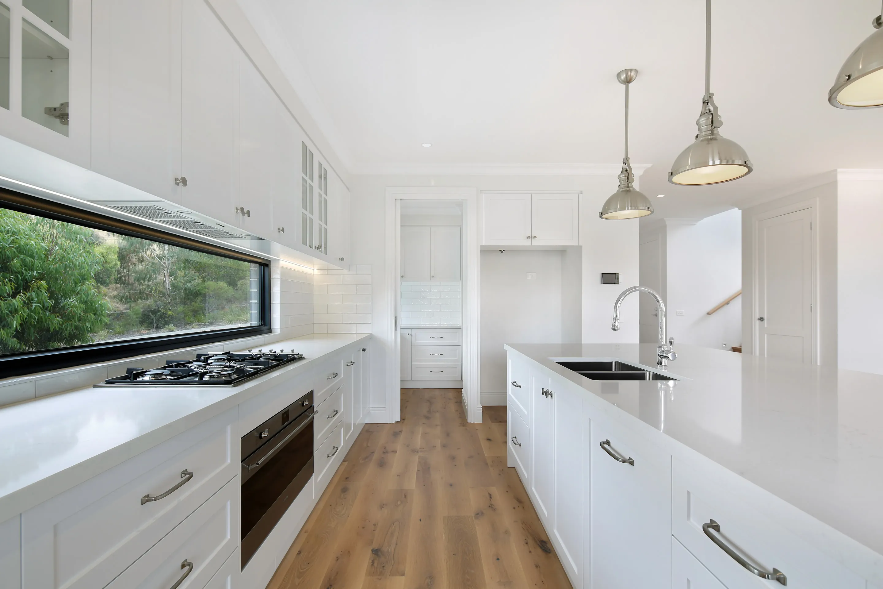 Galley kitchen with a gas cooktop and oven beneath a long window, a white island with sink and chrome tap, a butler's pantry beyond and oak floors