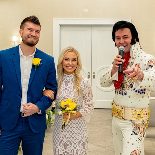 一对夫妻微笑地站在一位拿着麦克风的埃尔维斯·普雷斯利模仿者旁边。男士穿着蓝色西装,女士手中捧着一束花。