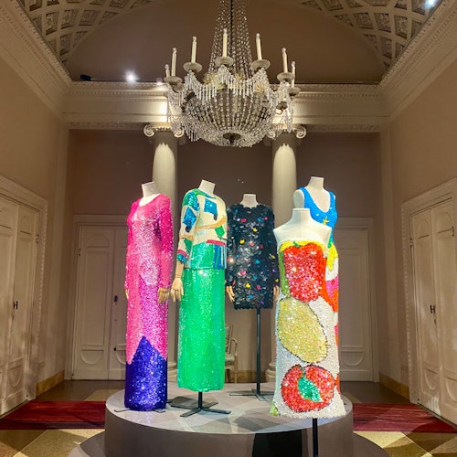 Four mannequins display sequined dresses in various colors under a large chandelier in an ornate room.