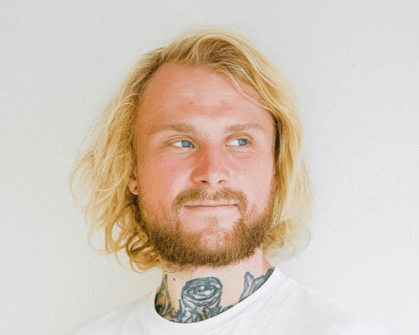 A white man with mid-length blonde hair and a beard stands against a white background. He is facing towards the camera, but looking off to the right.