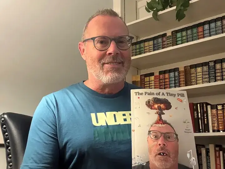 Stu holding a copy of his book “The Pain of a Tiny Pill: Finding My Way Back” in front of a bookshelf