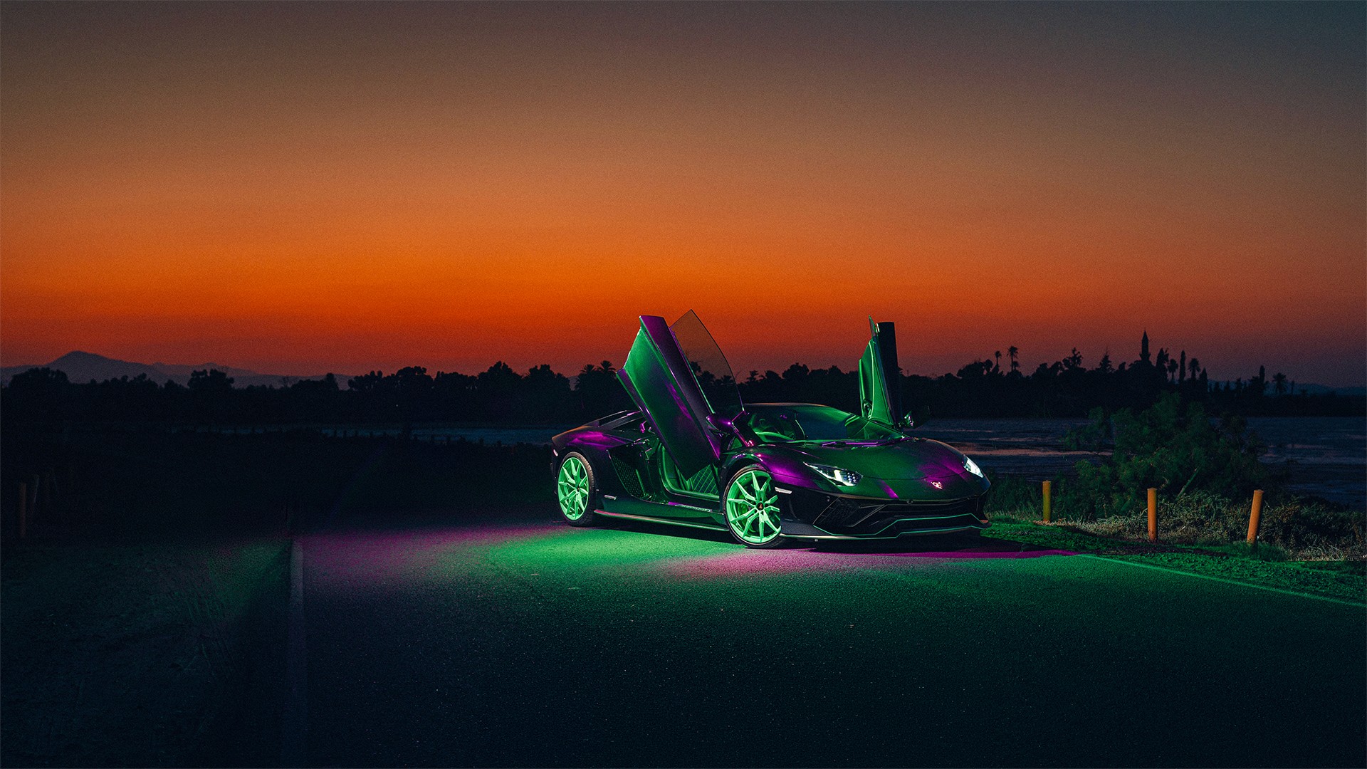 Lamborghini Aventador with both scissor doors raised, front three-quarter view against a deep orange sunset, the car and ground washed in green and magenta neon, with a distant tree-lined coast behind.