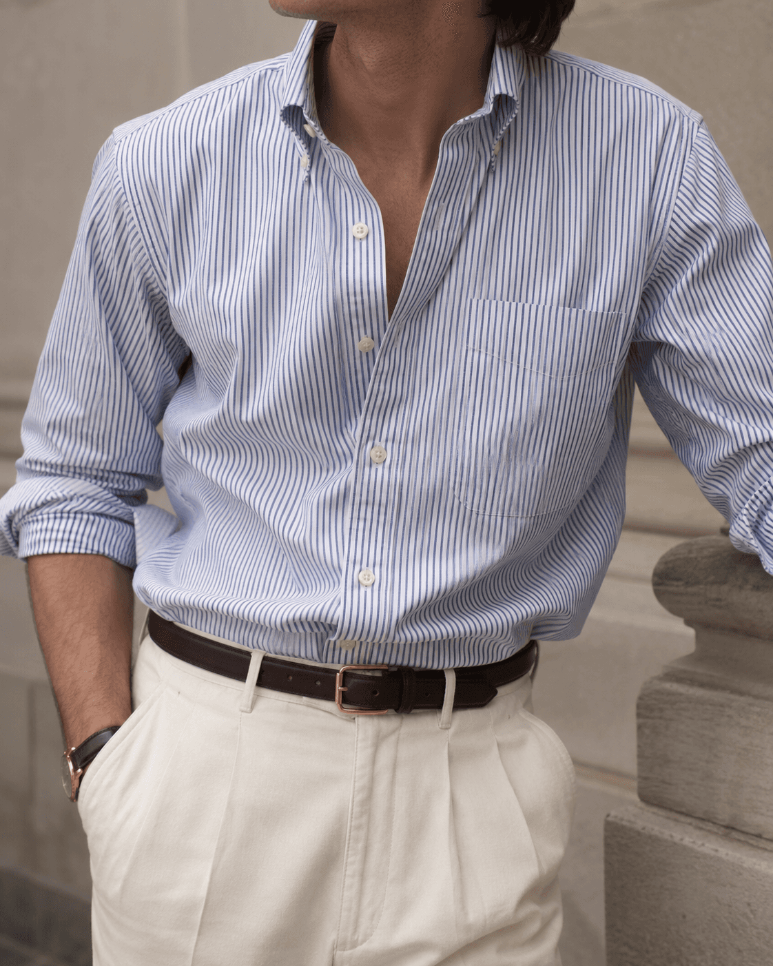 Close-up of a man in men's smart casual summer fashion, featuring a light blue and white striped cotton oxford cloth button-down shirt with rolled-up sleeves, tucked into high-waisted cream pleated linen trousers secured with a slim dark brown leather belt with a gold buckle.