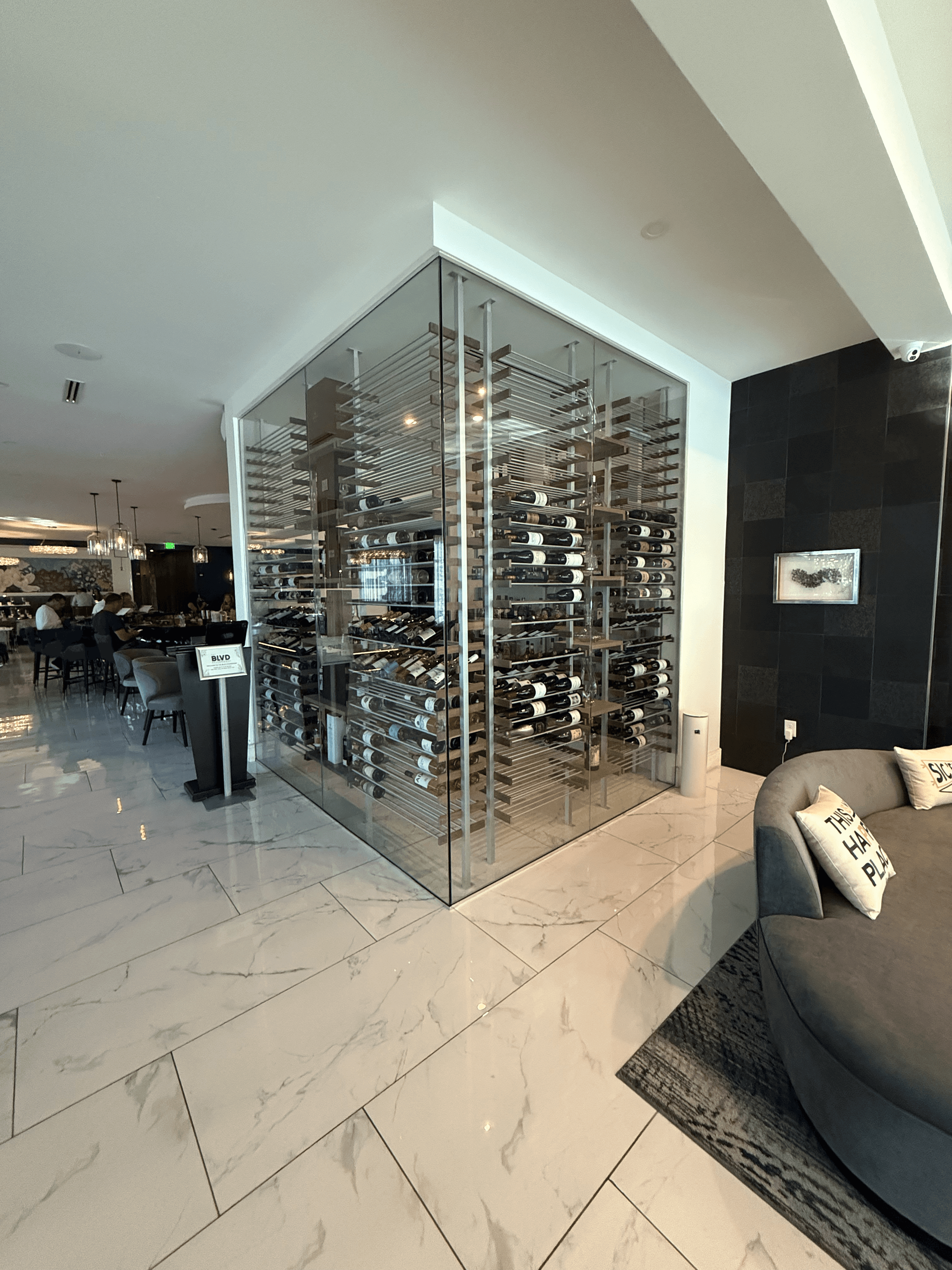 Modern glass-enclosed walk-in wine cellar at Cambria Waco Hotel featuring ½-inch frameless glass, aluminum and walnut racking, holding 880 bottles, designed and built by Prestige Wine Cellars.
