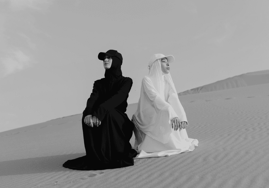 Two people wearing contrasting black and white traditional long robes and baseball caps are sitting back-to-back on a sandy dune.