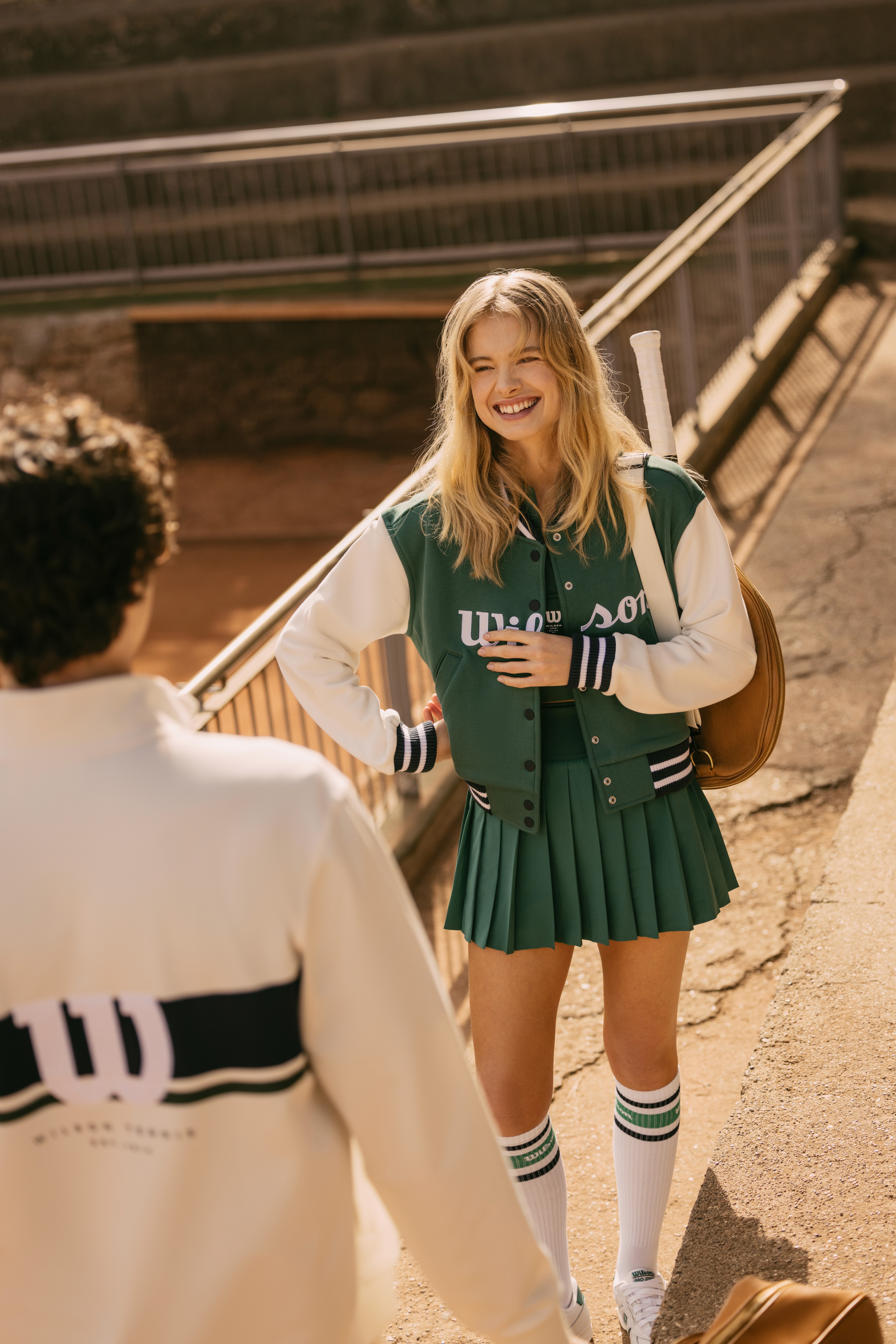 Une fille et un garçon vêtu de Wilson Courtstyle discute sur le court de tennis,avec jupe et blouson de la marque en blanc et vert