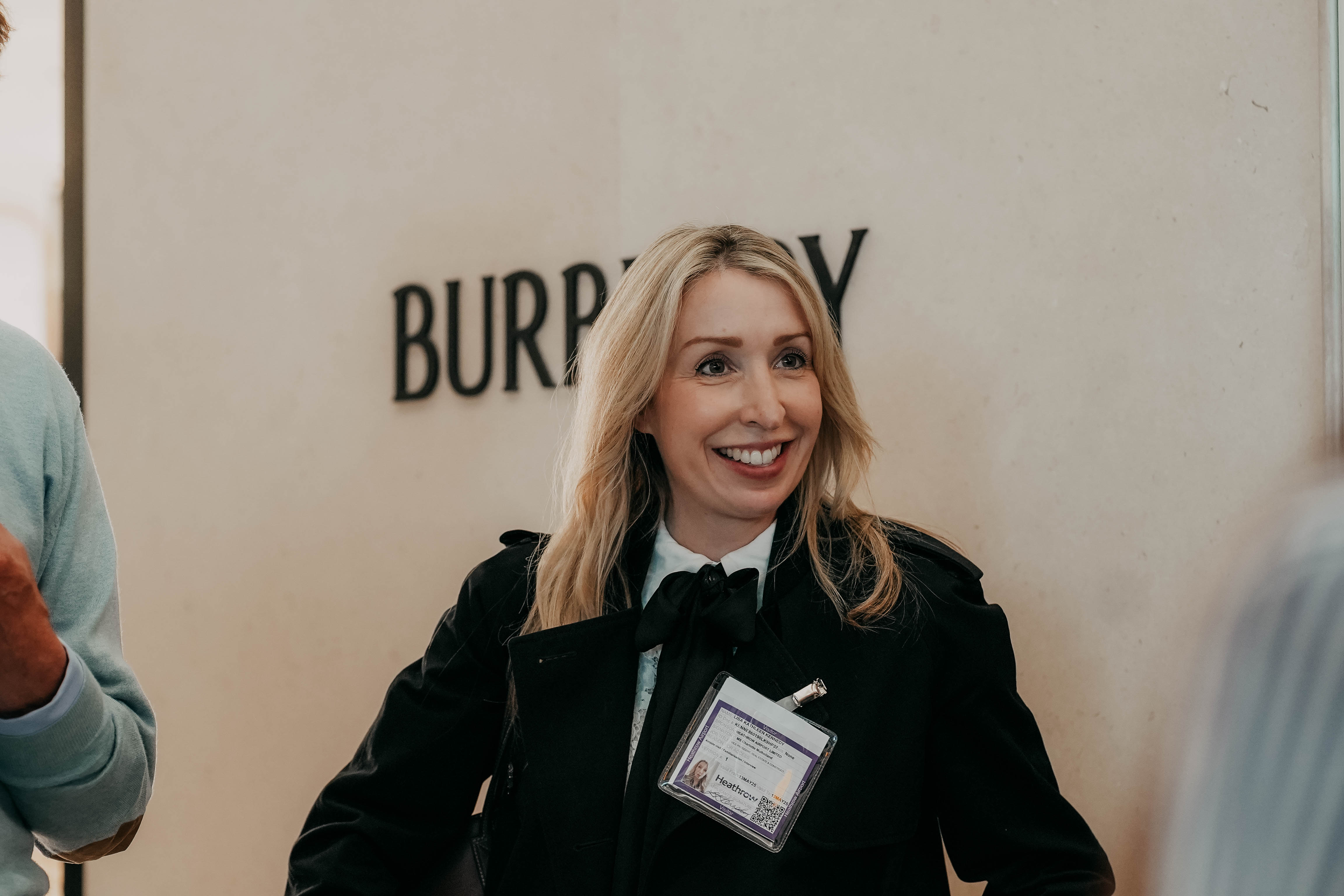 A blonde woman in a black jacket with a bow at the collar smiles as she looks to the side. She is standing in front of a wall with the Burberry logo and wearing a Heathrow event badge.