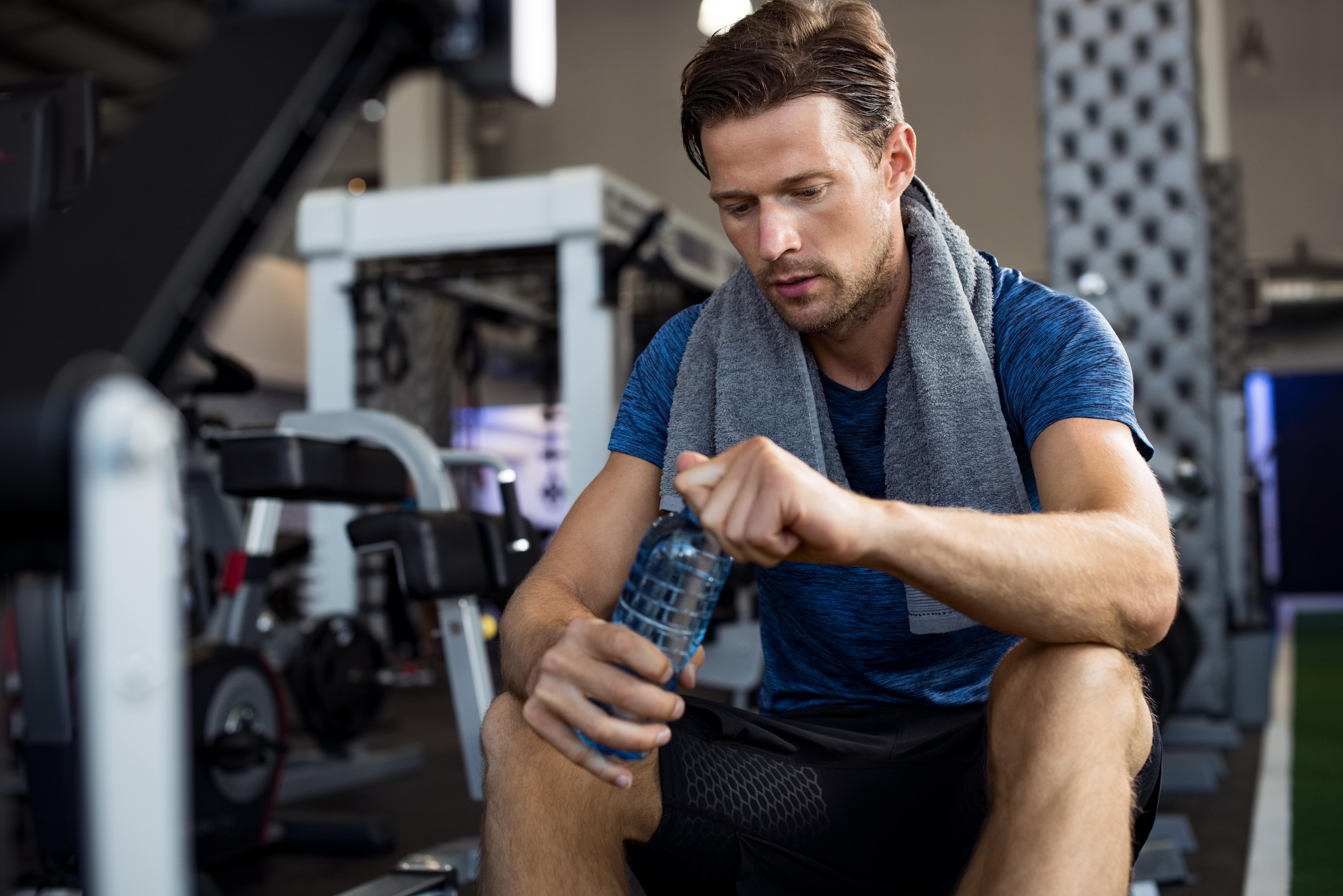 Man sitting in a gym, opening a water bottle with a towel around his neck, taking a break after a workout