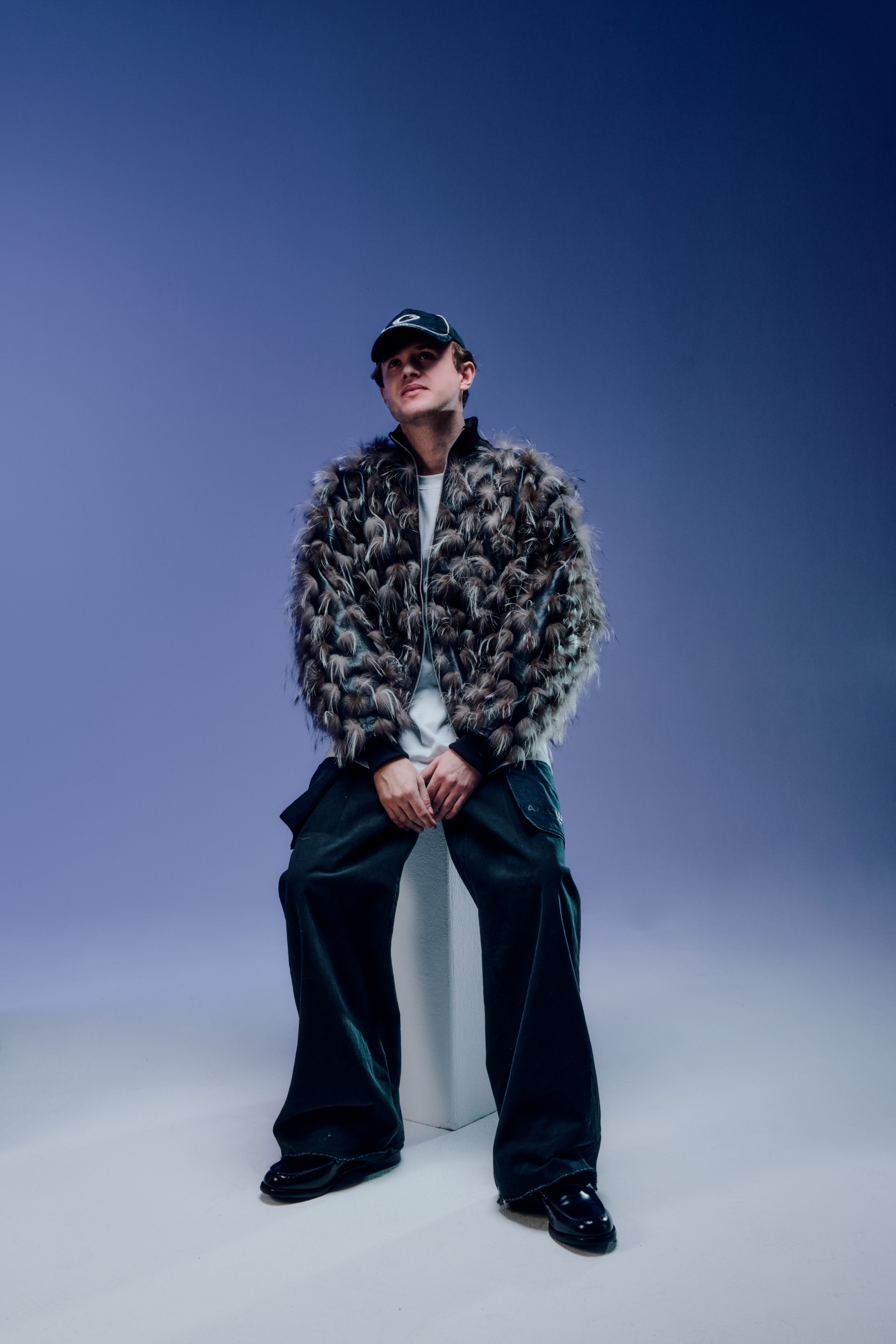 Marten Lou assis, portant une veste en fourrure texturée et une casquette, posant dans un style urbain et moderne.