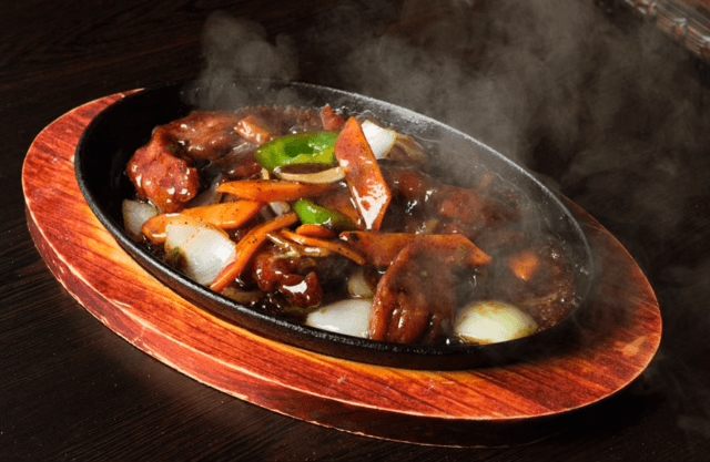 A steaming plate of sizzling beef stir-fry served on a hot iron platter, loaded with tender beef, bell peppers, carrots, and onions, coated in a savory glaze at Shanghai Times.