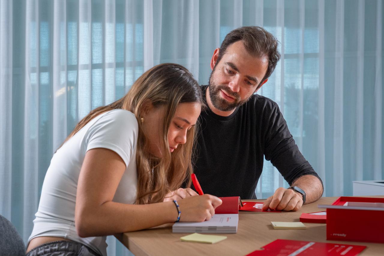 Two people sitting at a table, engaged in conversation while writing on paper, with a warm and friendly atmosphere. From KICKBOX program