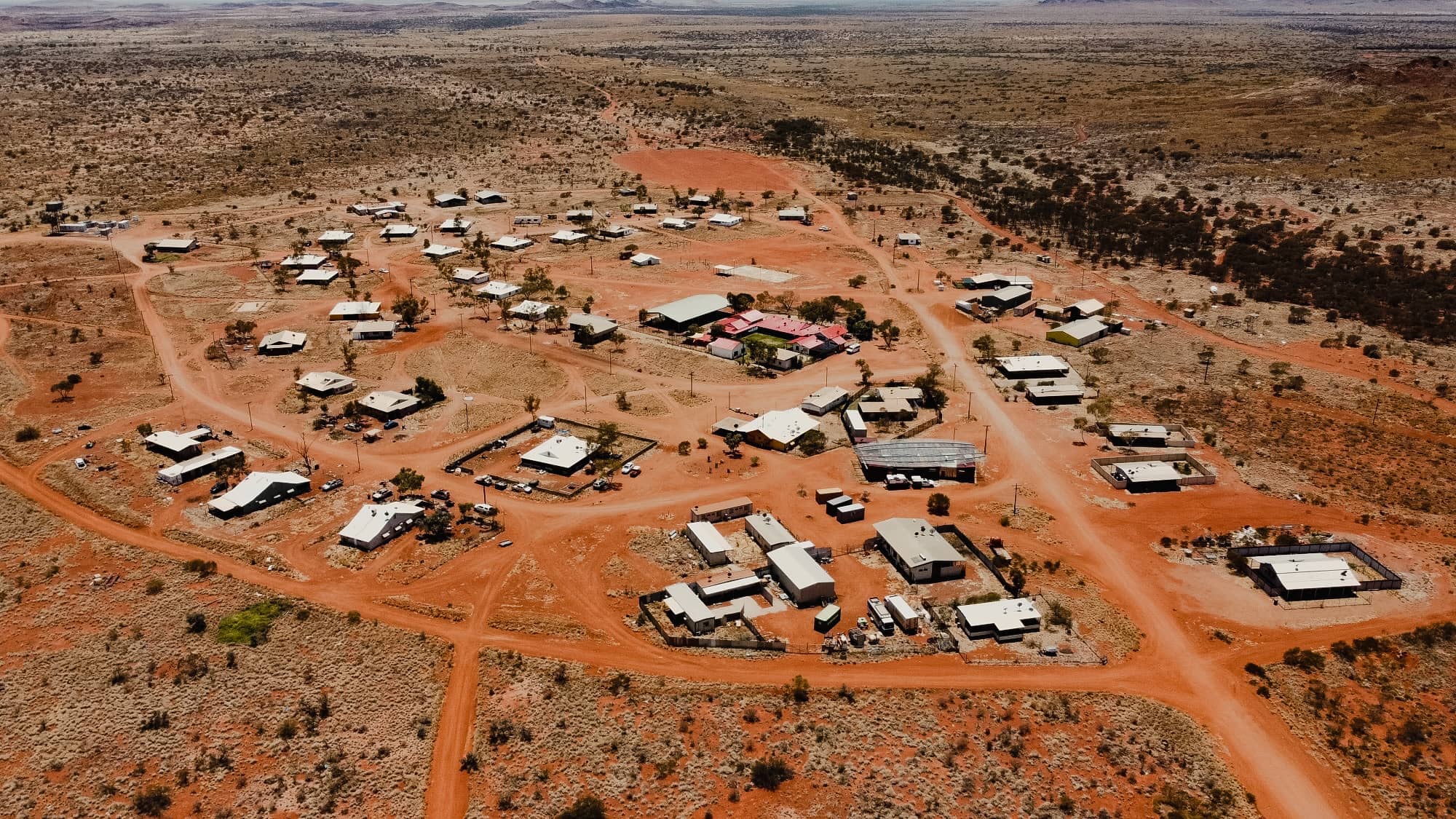 Aerial view of a remote Australian outback community with red dirt roads and scattered buildings under a hazy sky.