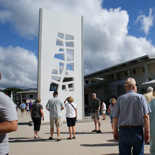 ผู้คนเดินอยู่ใกล้ประติมากรรมสีขาวสูงที่มีลวดลายตัดแบบนามธรรม ภายใต้ท้องฟ้าที่มีเมฆเป็นบางส่วน ในบรรยากาศของลานกลางแจ้ง