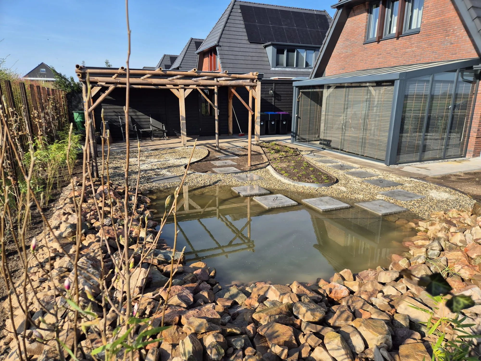 Japanse tuin met vijver omringd door rotsen, houten pergola, grindpad met stapstenen en nieuwbouwwoning in Gorredijk.