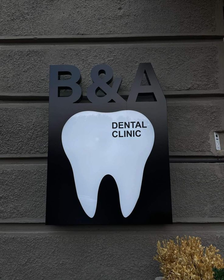 Exterior B&A Dental Clinic sign featuring a tooth-shaped illuminated logo mounted on a dark wall plaque.