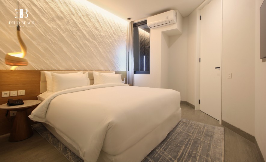 A cozy and minimalist bedroom at Echo Beach Suite in Canggu, Bali, with a soft white bed, a textured illuminated wall, air conditioning, and warm décor for a tranquil accommodation experience close to Echo Beach.