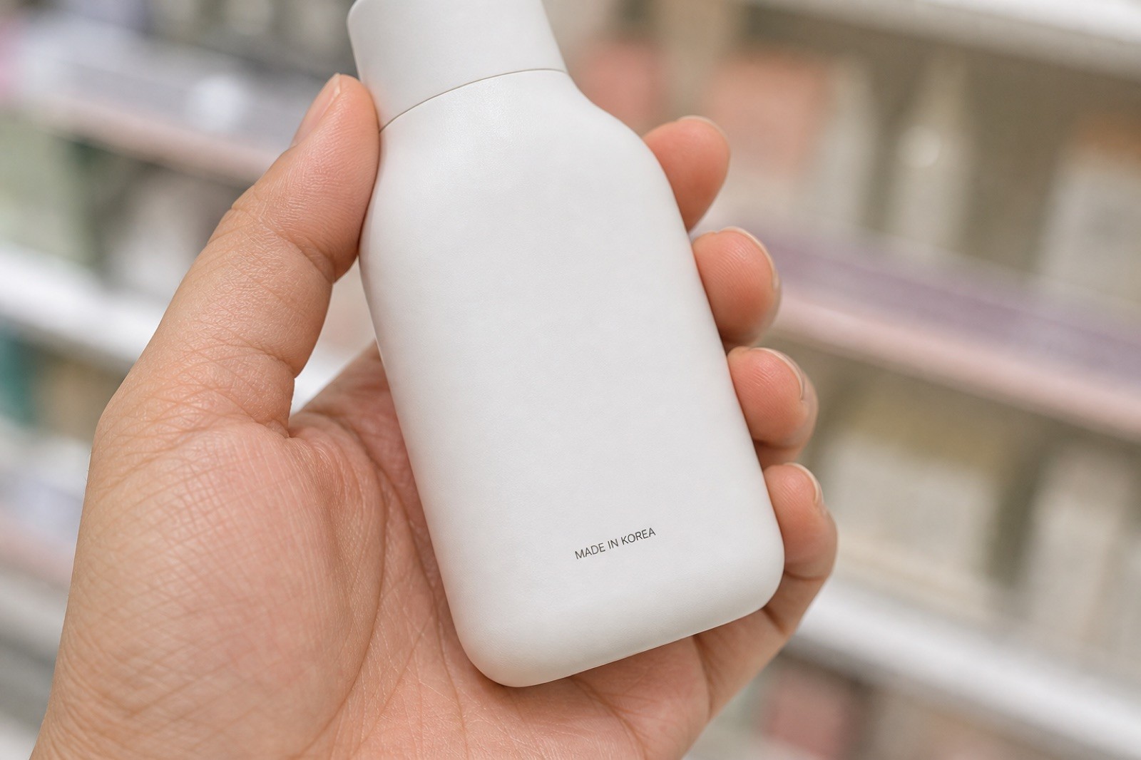 Close-up of a hand holding an unbranded white skincare bottle, with 'MADE IN KOREA' printed as the only text on the package